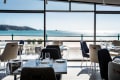 Dining room with upholstered chairs and wooden tables in the foreground, and a floor-to-ceiling panoramic window looking out to a sandy beach and blue sea