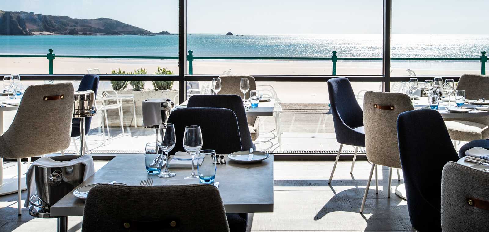 Dining room with upholstered chairs and wooden tables in the foreground, and a floor-to-ceiling panoramic window looking out to a sandy beach and blue sea