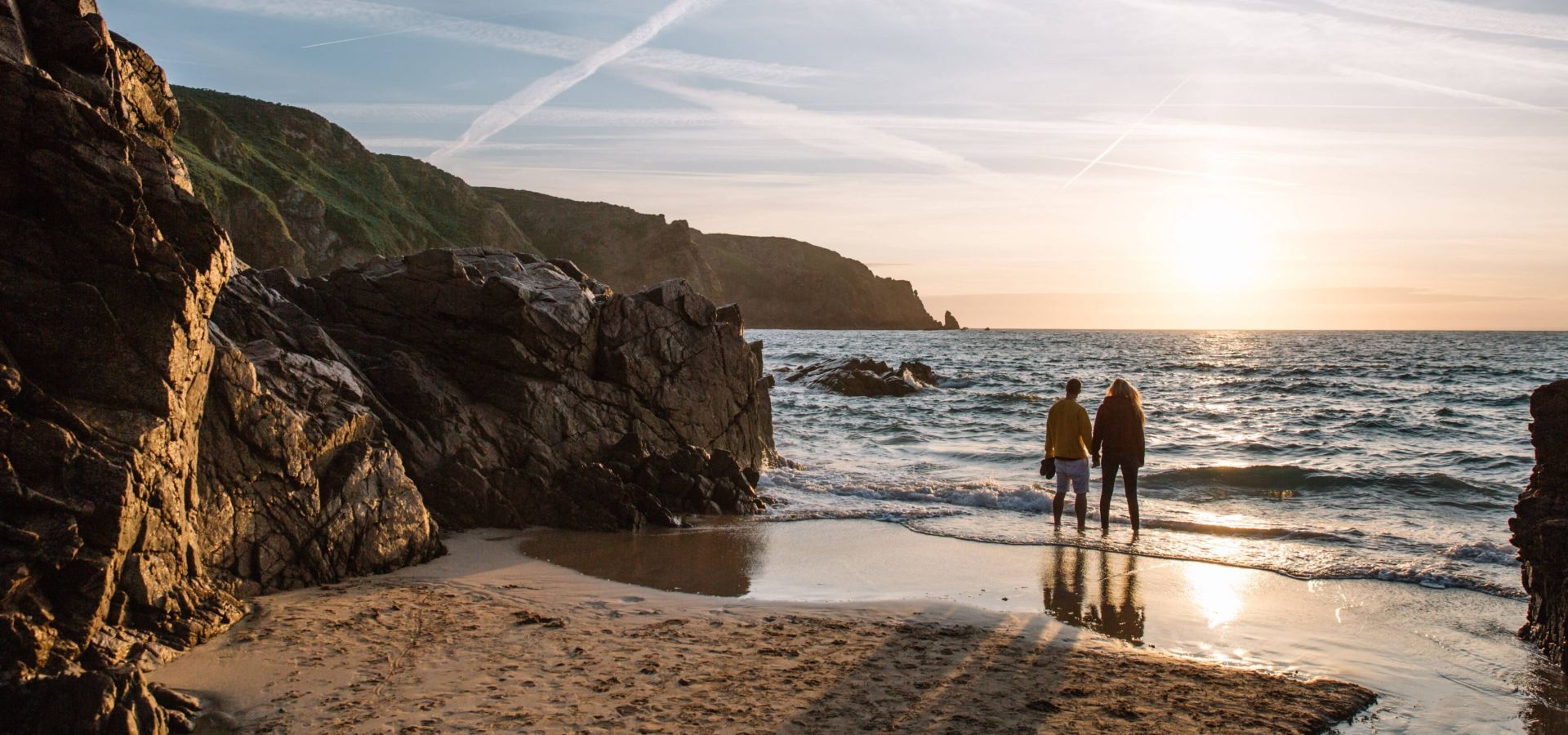 A couple stand at the shoreline as they admire the sunset over Plu00e9mont Bay