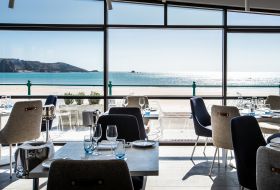 Dining room with upholstered chairs and wooden tables in the foreground, and a floor-to-ceiling panoramic window looking out to a sandy beach and blue sea