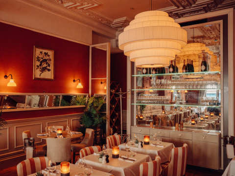 A wide shot of the dining room, including a dark rust-coloured feature wall, framed portrait paintings, large light fixture, and Victorian fireplace