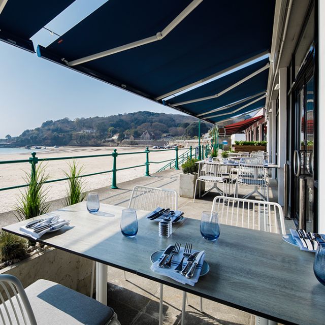 Tables under a blue awning, overlooking the beach at St Brelade