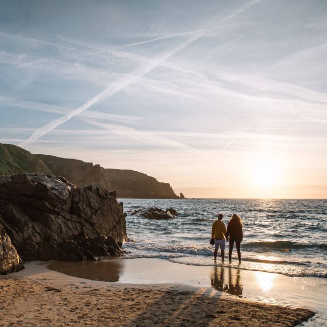 A couple stand at the shoreline as they admire the sunset over Plu00e9mont Bay