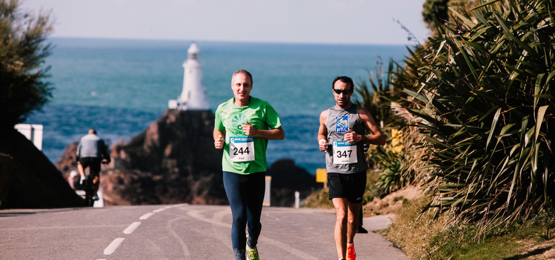 Two men running in the Jersey Marathon with Corbiere Lighthouse behind them