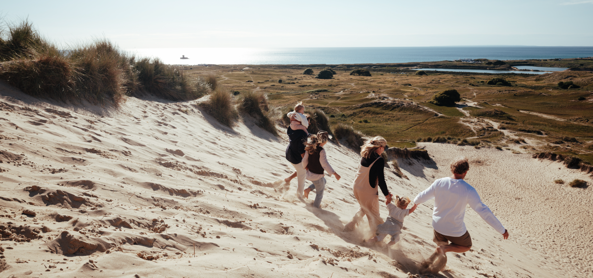 Family running on sand dunes in the sunshine with sea in the background