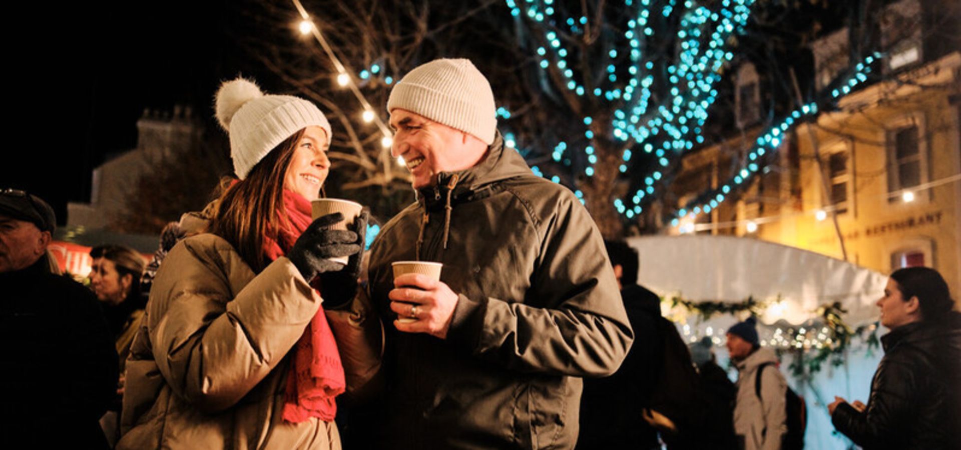 Couple smiling each other. They are wearing warm, wooly, coats are are surrounded by people and bright Christmas lights.