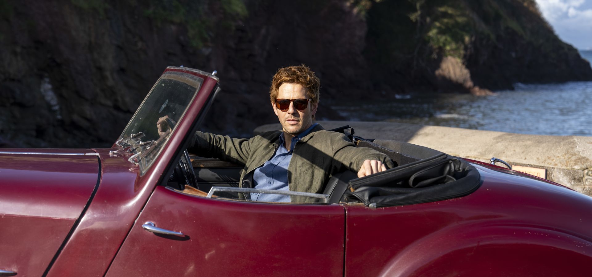 An image of Damien Molony, in character as Bergerac sat inside the iconic, 1947 Triumph Roadster.