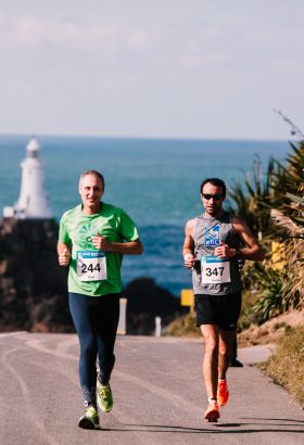 Two men running in the Jersey Marathon with Corbiere Lighthouse behind them