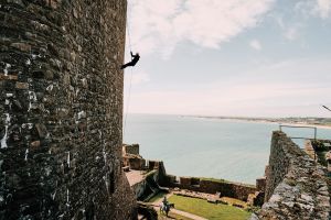 A person abseiling down a medieval castle. The castle is overlooking a large bay.