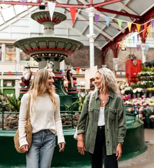 Two women in a victorian market, with a large central fountain behind them.