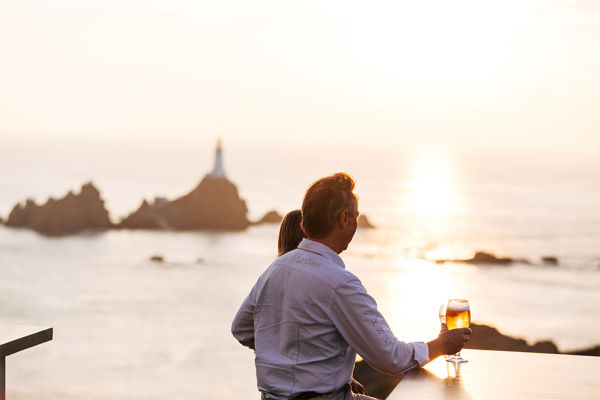 A couple sit looking out to La Corbiere Lighthouse at sunset having a drink at Corbiere Phare