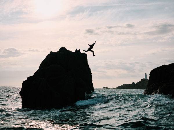 A silhouette of a man rock jumping into a sea between to rocks. The water is choppy and it is a sunny day.