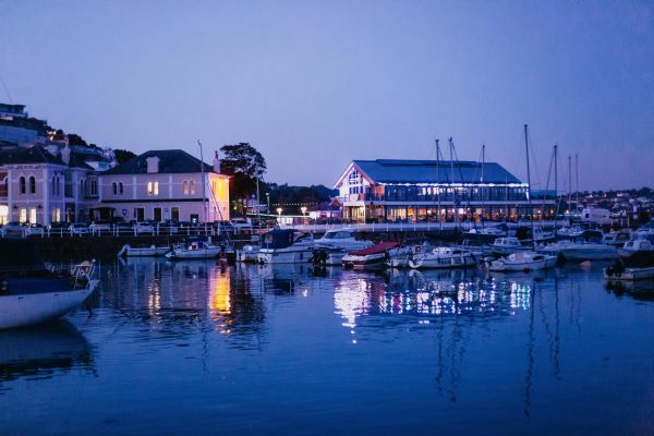 A small harbour town at dusk named St. Aubin. There are a few boats moored in the Marina, and a restaurant lit up by bright lights.