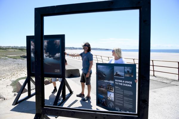Man and a a woman standing in front of the sea at next to an exhibition of giant photographs
