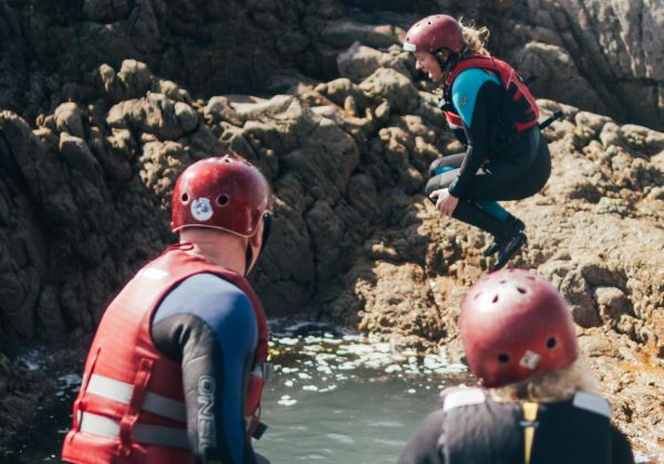 People coasteering in Jersey waring wetsuits, old trainers and a hard hat. There is a girl jumping into a rock pool.