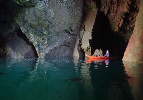A man in a Kayak exploring a dark sea cave. There is light coming the the mouth of the save cave, lighting the surface of the water and his kayak.