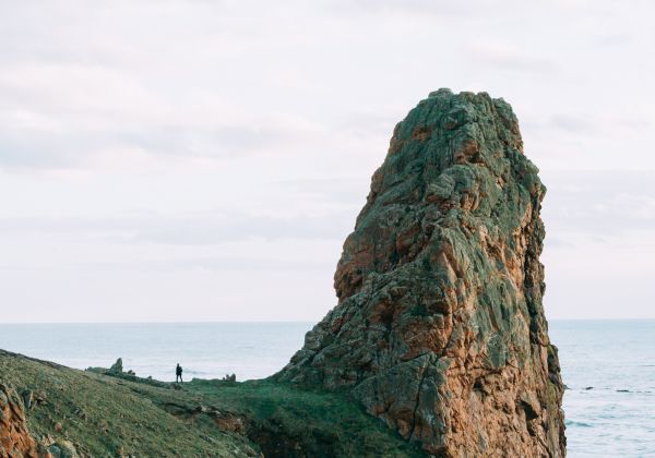 A large rocky formation rising out of the edge of a cliff. There is a man standing at the base of the formation.