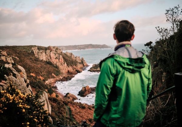 A man in a green raincoat looks out along a rugged coastline in autumn